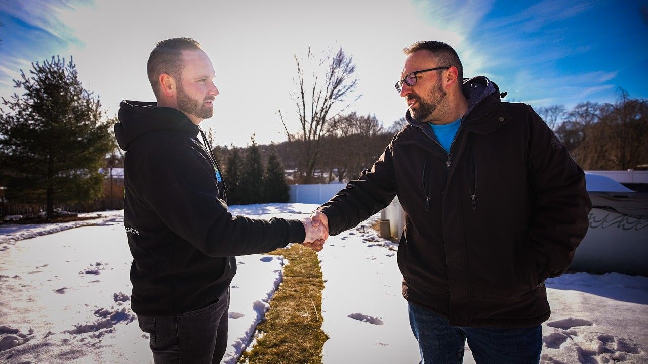 Two men shaking hands outdoors on a snowy path under a bright blue sky.