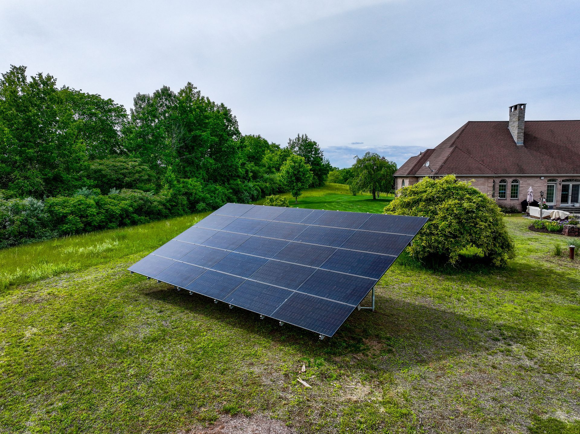 Solar panel array in a grassy yard beside a house and trees under a cloudy sky