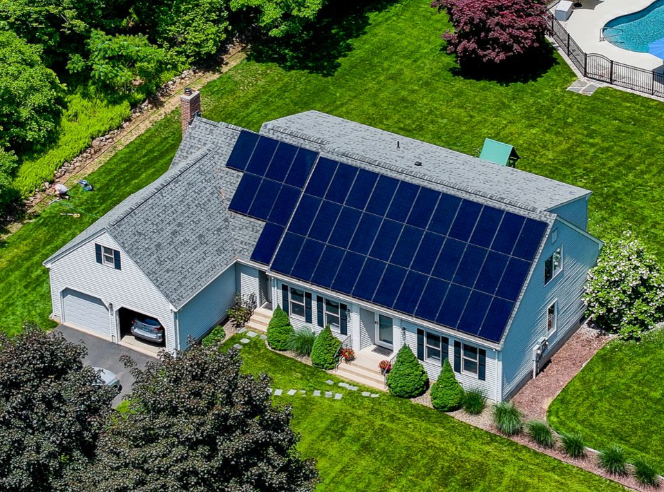 Aerial view of a white house with a large rooftop solar panel array and green lawn.