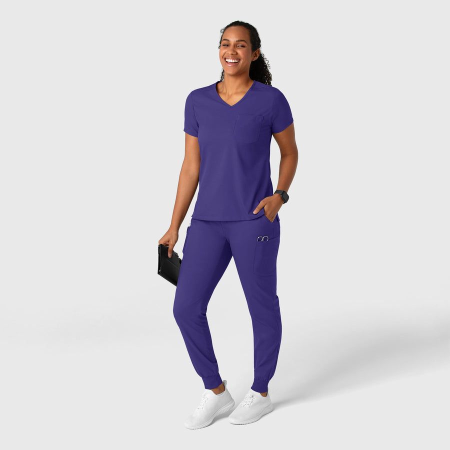 Woman wearing purple scrubs smiles, holding a tablet, standing against a white backdrop.