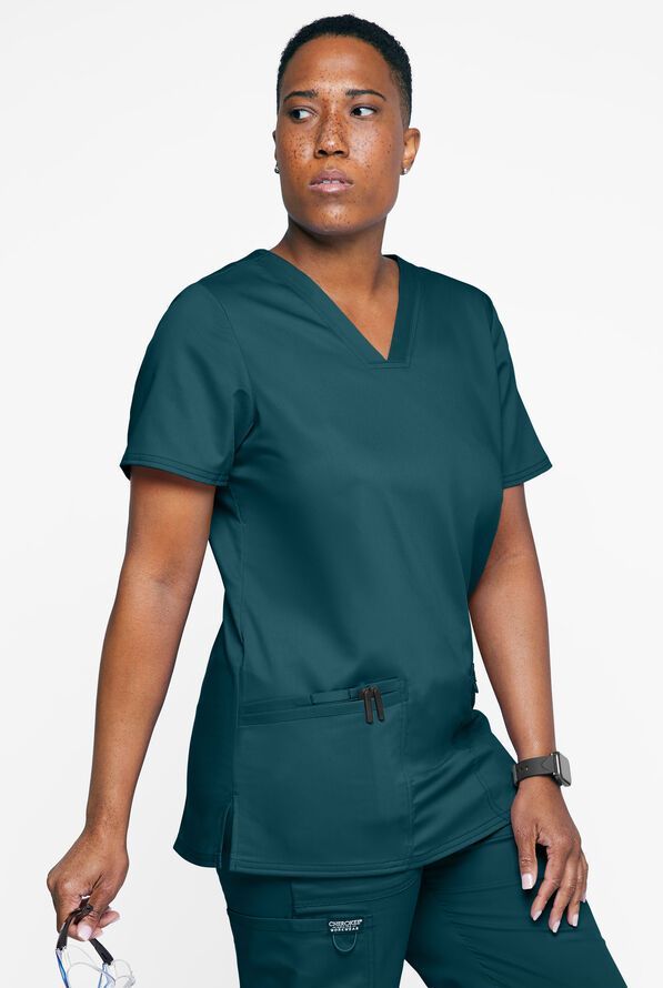 Person in teal scrubs, looking to the side, holding glasses, indoors.