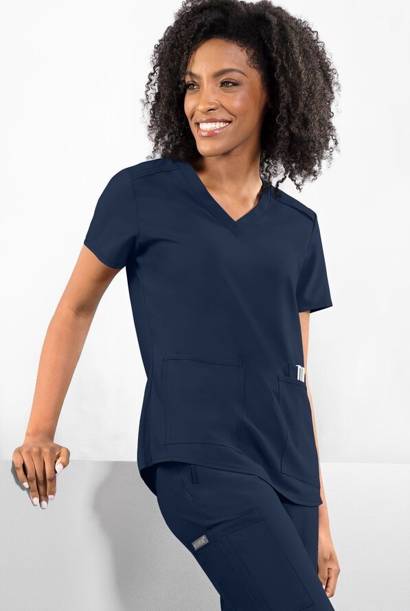Woman in navy scrubs, smiling, leaning against a white surface, in a studio setting.