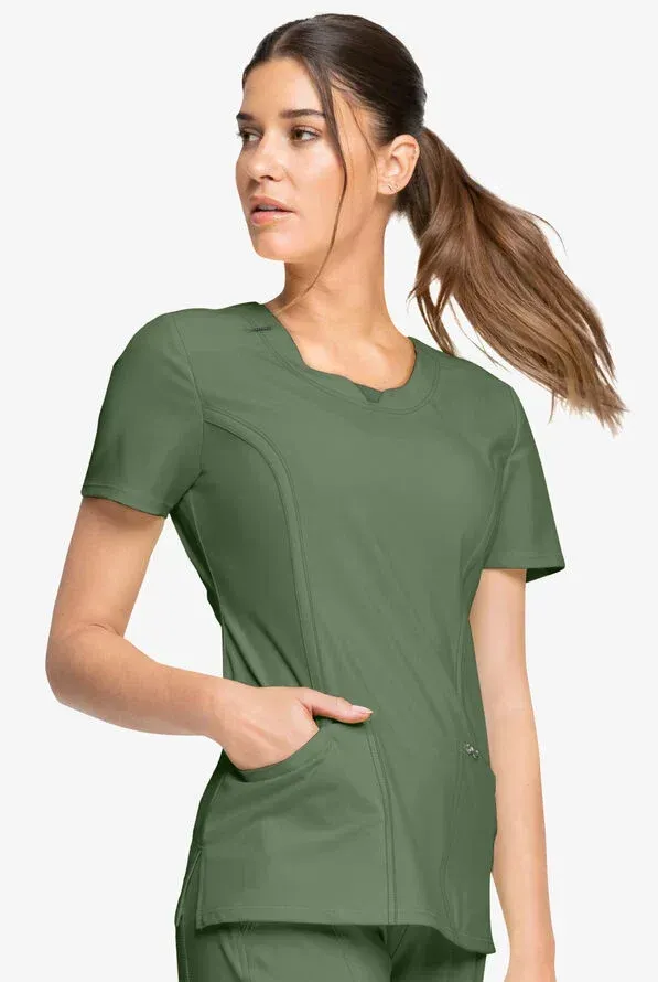 Woman in green scrubs with a unique neckline, hand in pocket, looking up.
