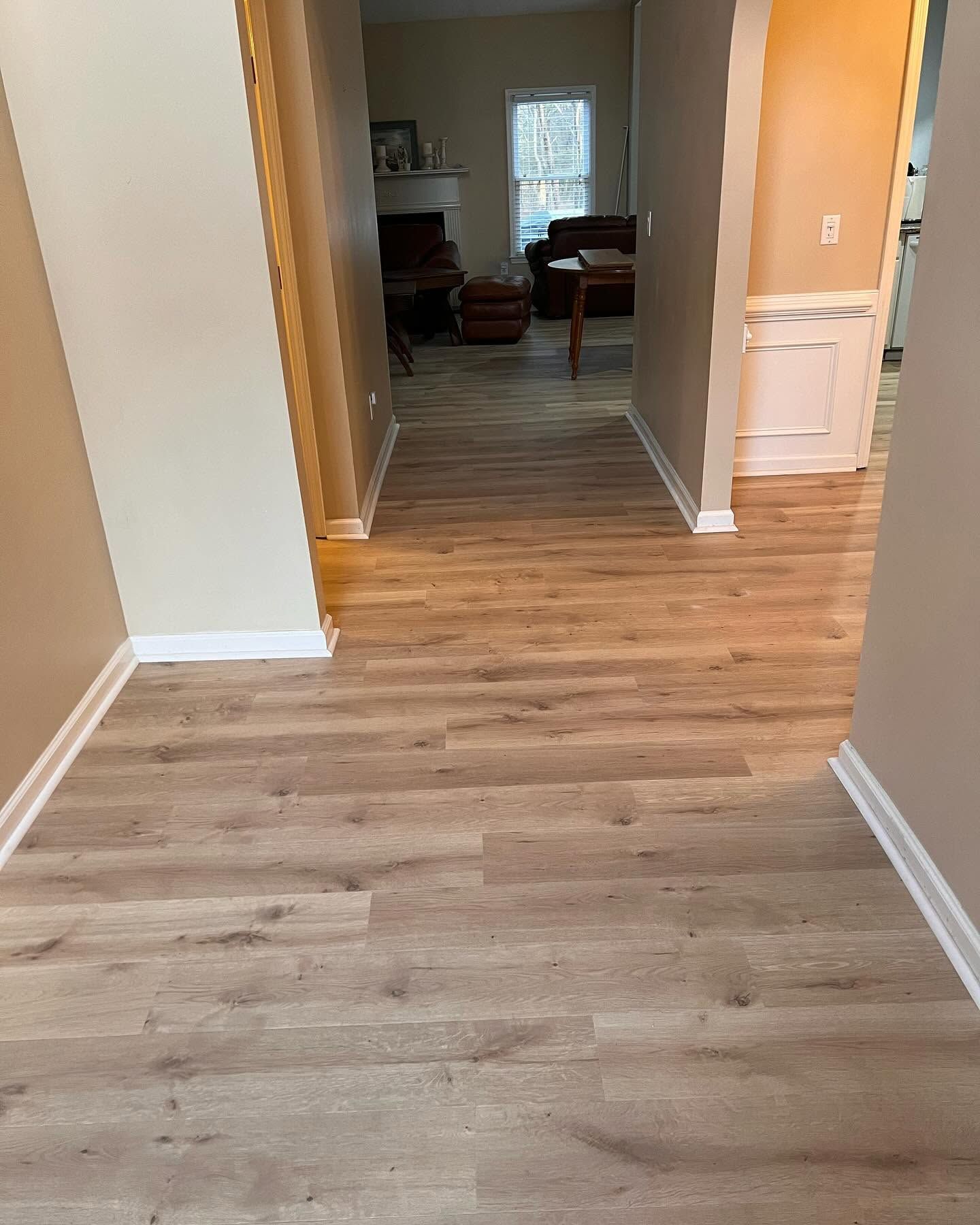 Hallway with light wood flooring leading to a living room, beige walls, and white trim.