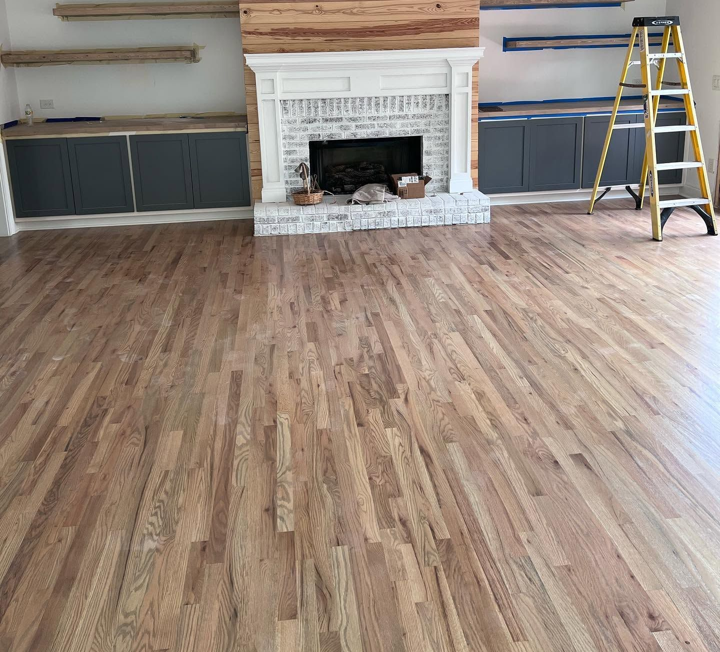 Empty living room with hardwood floors, white fireplace, gray built-ins, and a yellow ladder on the right.