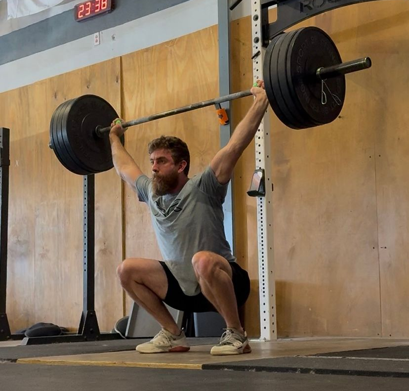 Man in pink shorts overhead squats with barbell in gym.