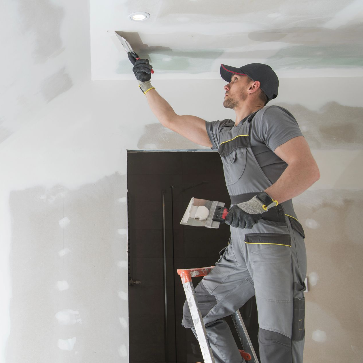 Persona su una scala che applica il composto per cartongesso al soffitto con una cazzuola.