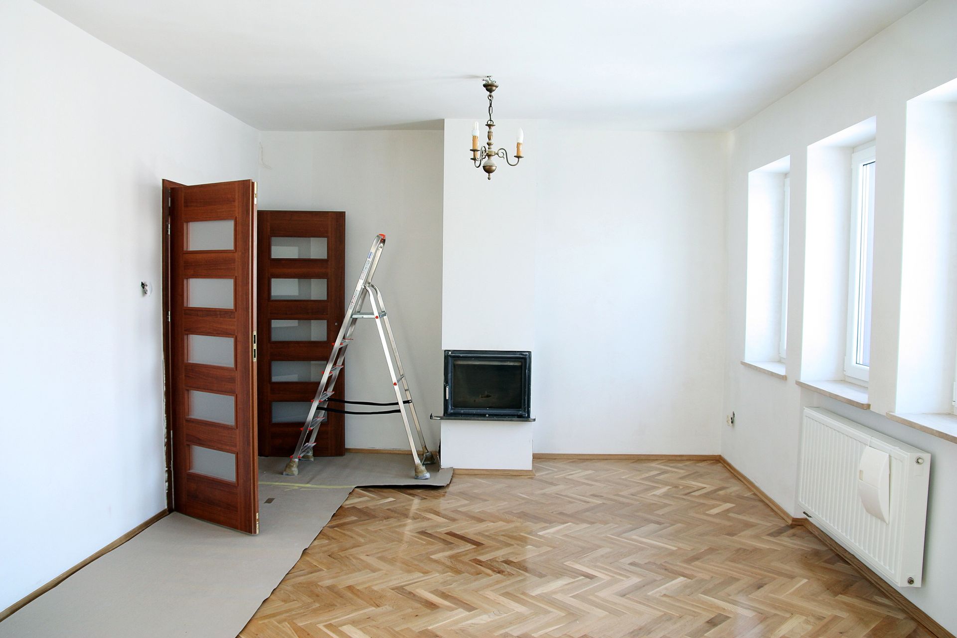 Stanza vuota con pavimenti in legno, camino e due porte appoggiate a un muro; lì vicino c'è una scala.