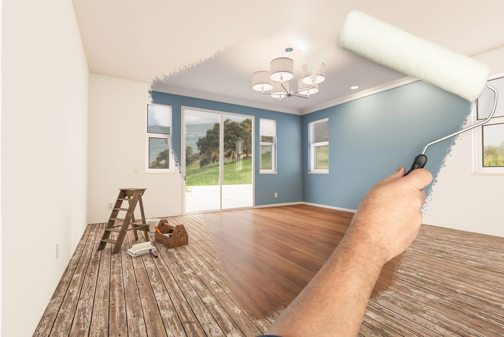 Una mano che usa un rullo di vernice trasforma i colori di una stanza: pareti blu, soffitto bianco e pavimento in legno.