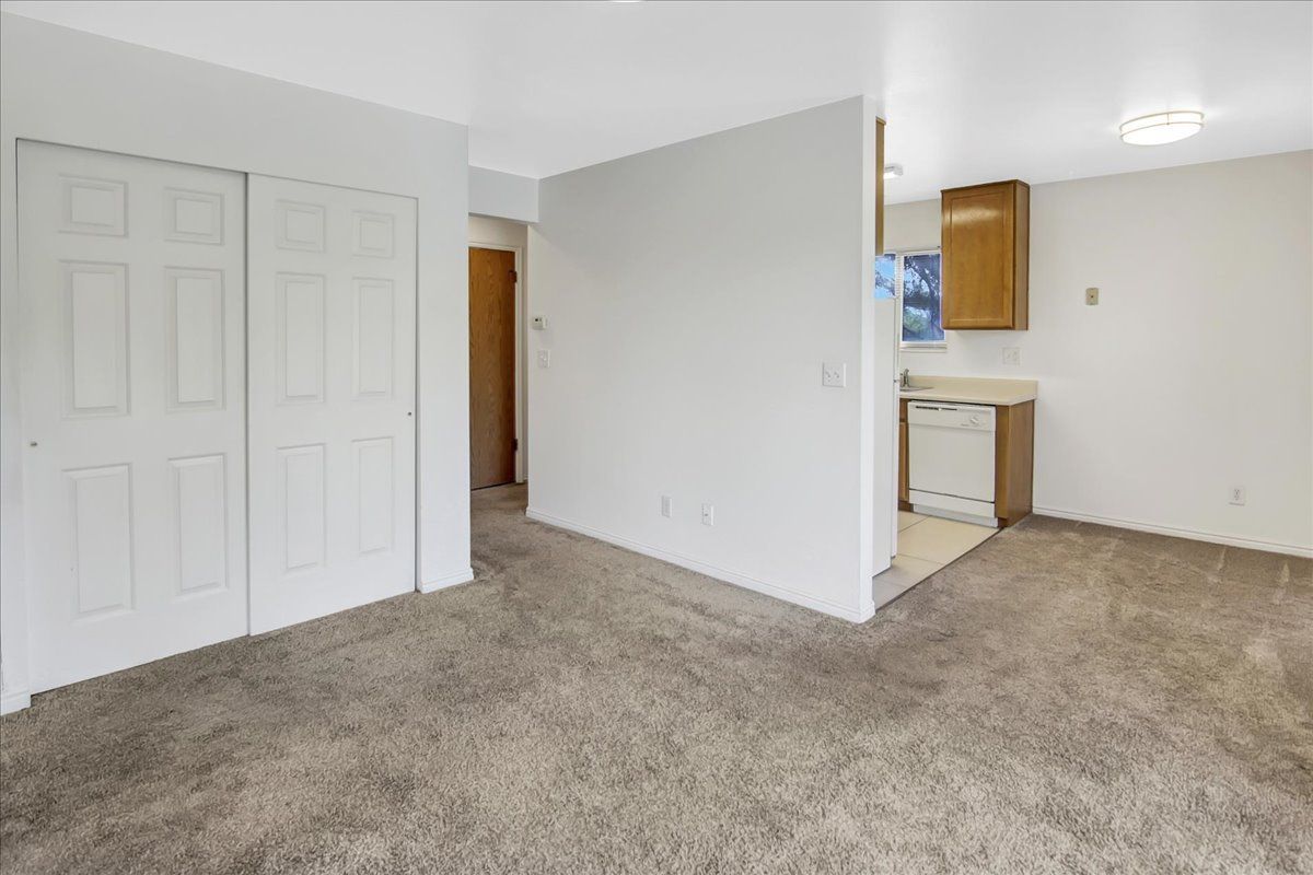 Empty living space with gray carpet, white walls, closet doors, and a partial view of the kitchen.