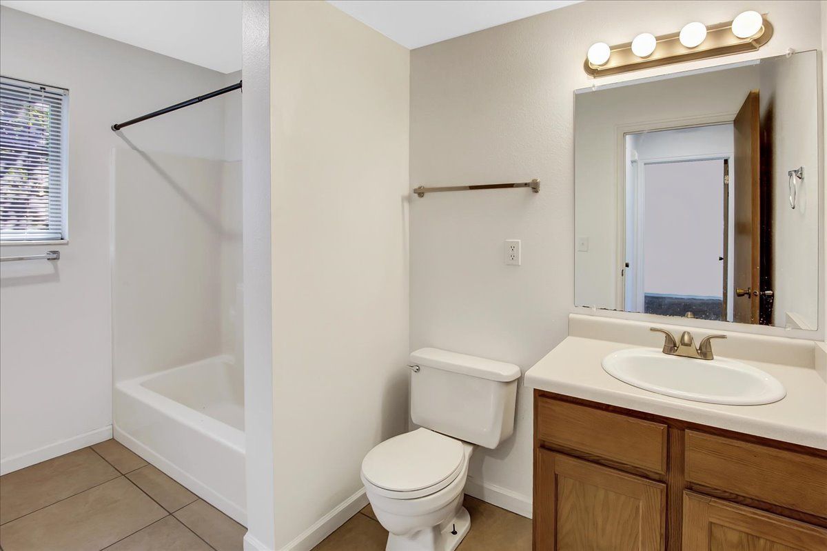 Bathroom with white tub, toilet, and vanity with mirror and lights; beige tile floor.