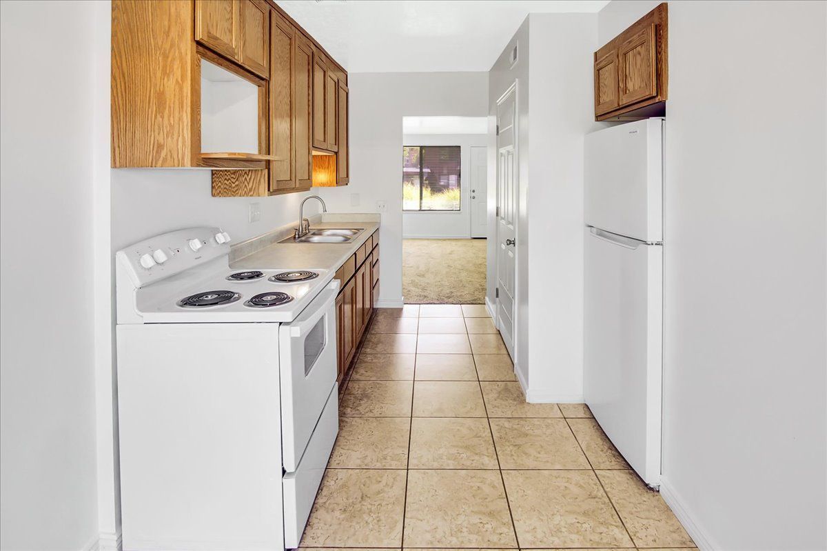 Small, galley-style kitchen with white appliances and light brown cabinets. Tile flooring leads to a hallway.