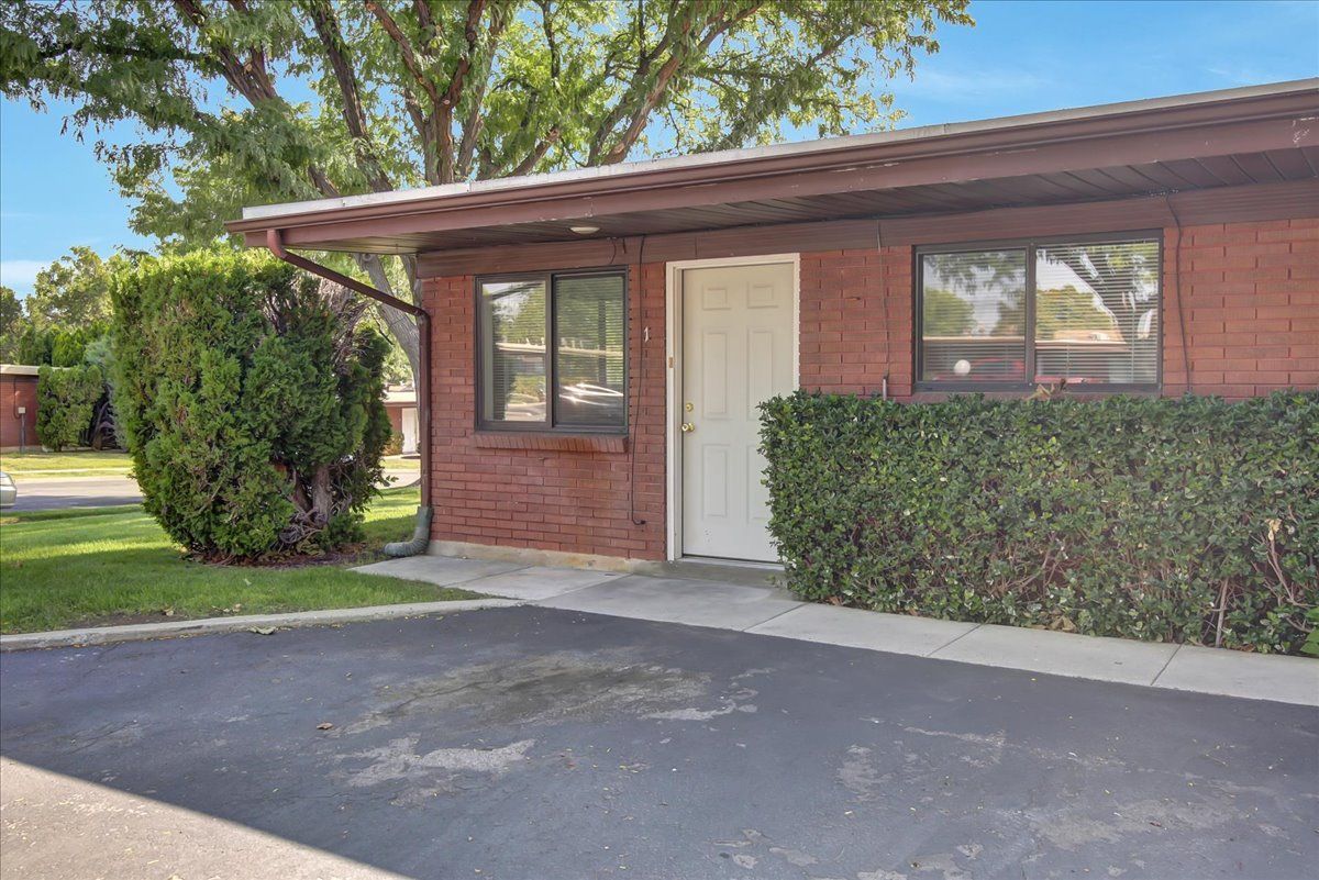 Brick building with a white door, two windows, and dark asphalt driveway, bushes and greenery surround it.