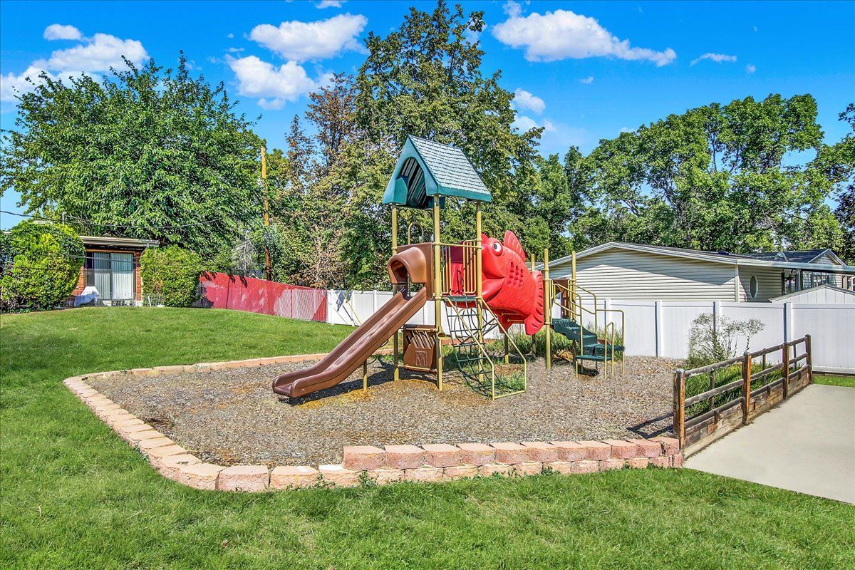 Playground with a slide, climbing structure, and wood chips, on a grassy lawn with a fence.