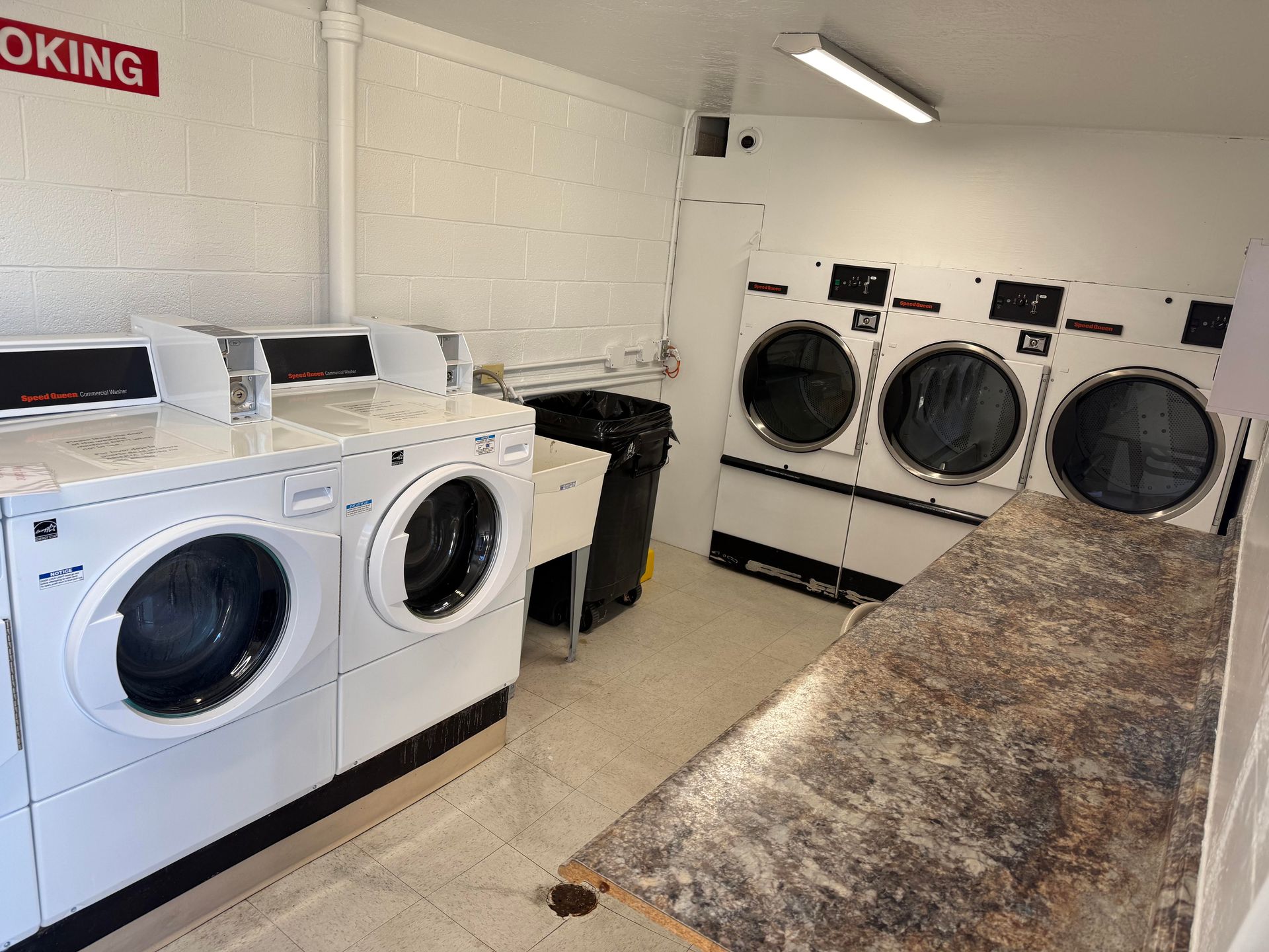 Laundry room with washers, dryers, folding table, and trash can. White walls, beige floor.