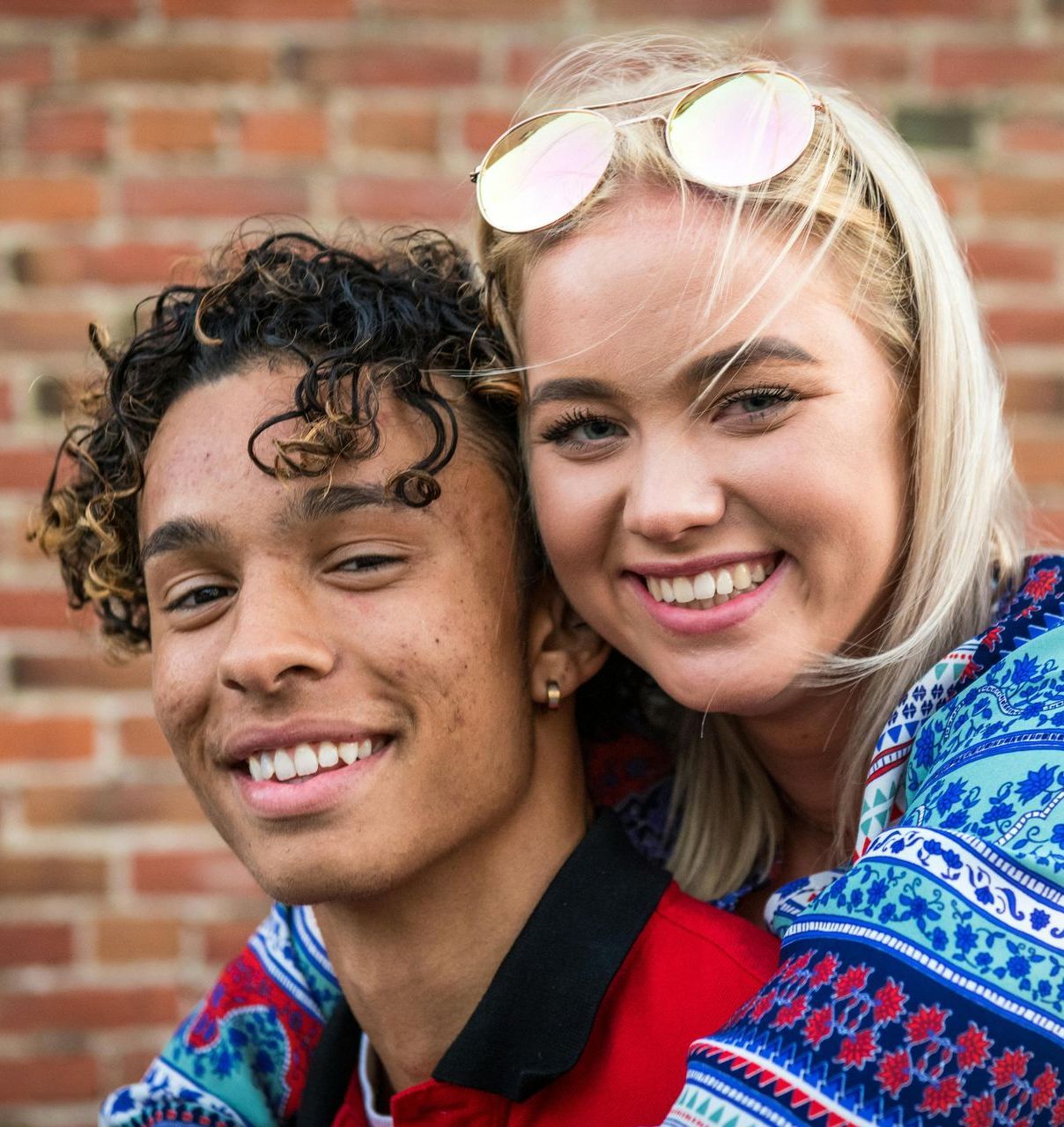 Smiling couple posing together in front of a brick wall; woman's arms around man.