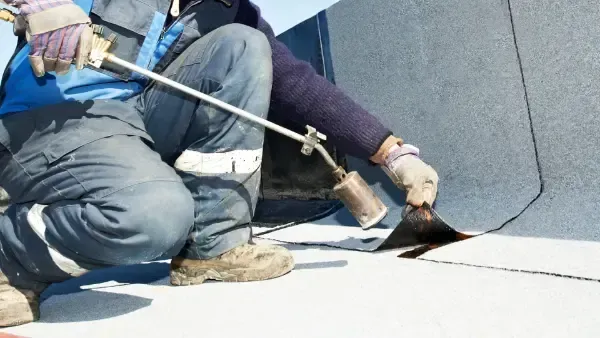 A man is kneeling down and using a torch to apply a roof coating.
