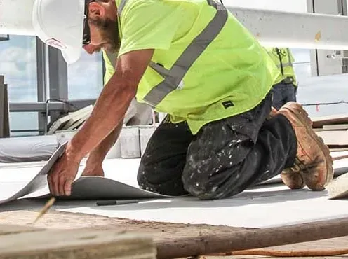 A construction worker is kneeling down on the ground while working on a roof.