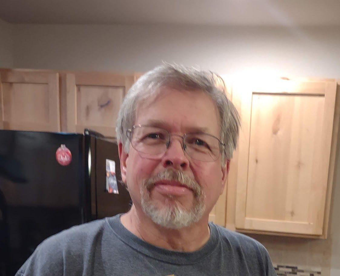 A person with glasses and a grey beard wearing a grey t-shirt in a kitchen.