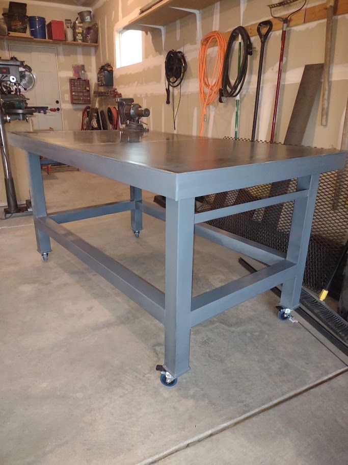 A sturdy, gray metal workbench with a smooth top sits on wheels in a garage, surrounded by tools and shelving.