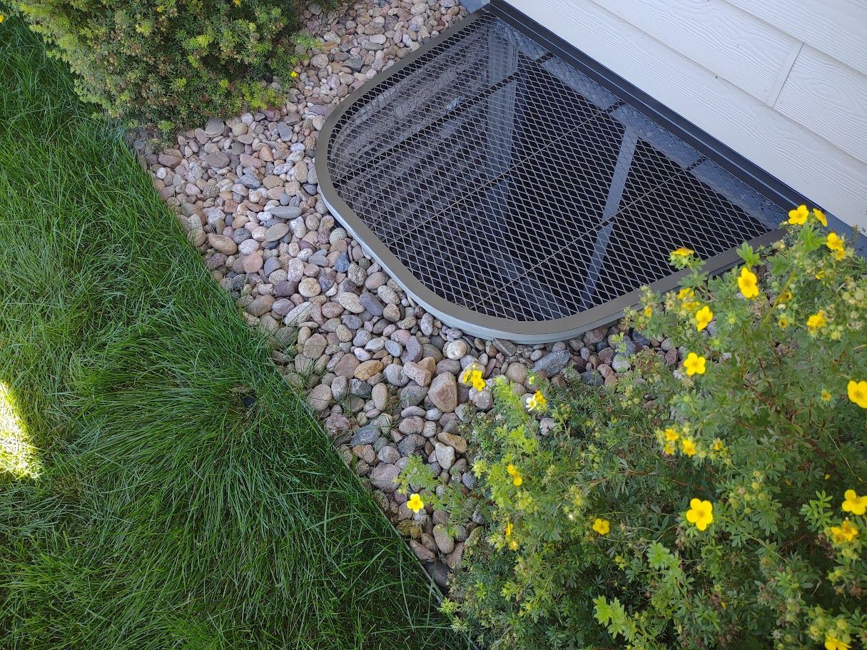 A black window well cover sits on a stone-filled garden bed next to a house wall, a green lawn, and a yellow-flowered shrub.
