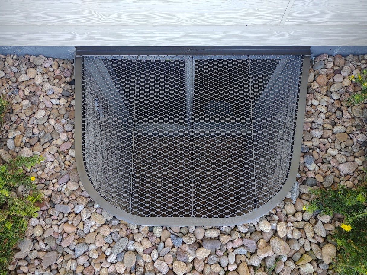 A black metal basement window well cover sits over a gravel-covered ground next to a home foundation.