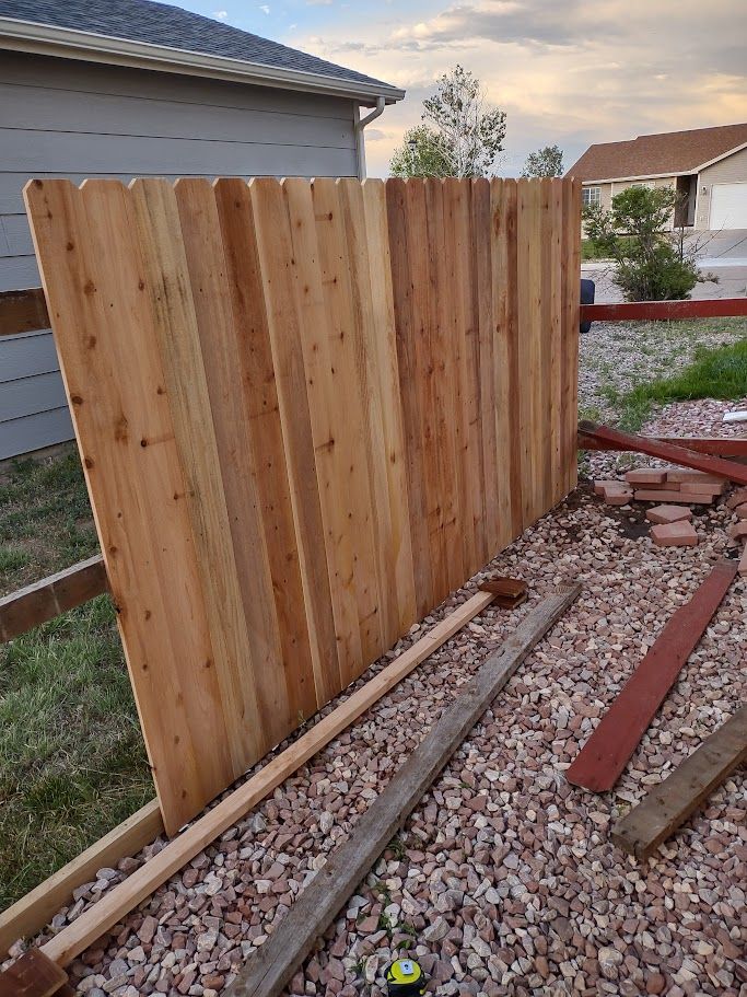 A section of a cedar wood fence under construction, showing vertical pickets against a house in a rocky yard.