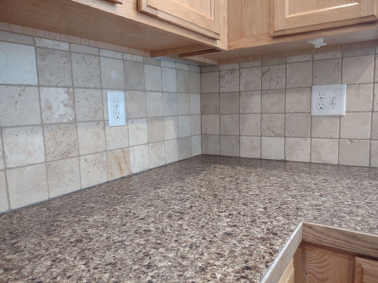 A kitchen corner with tan stone-look square tiles on the backsplash and a speckled brown granite countertop.