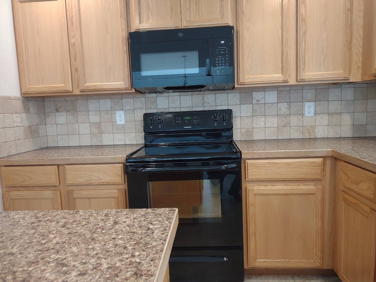 A kitchen featuring light wood cabinets, a black stove and microwave, and a textured countertop with stone-tiled backsplash.