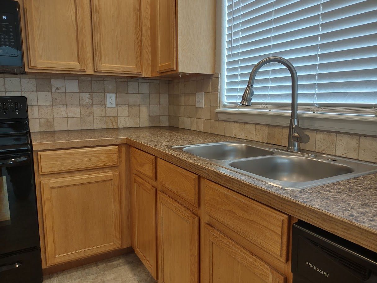 A kitchen counter with wood cabinets, tan tiled backsplash, a double stainless steel sink, and a high-neck faucet.