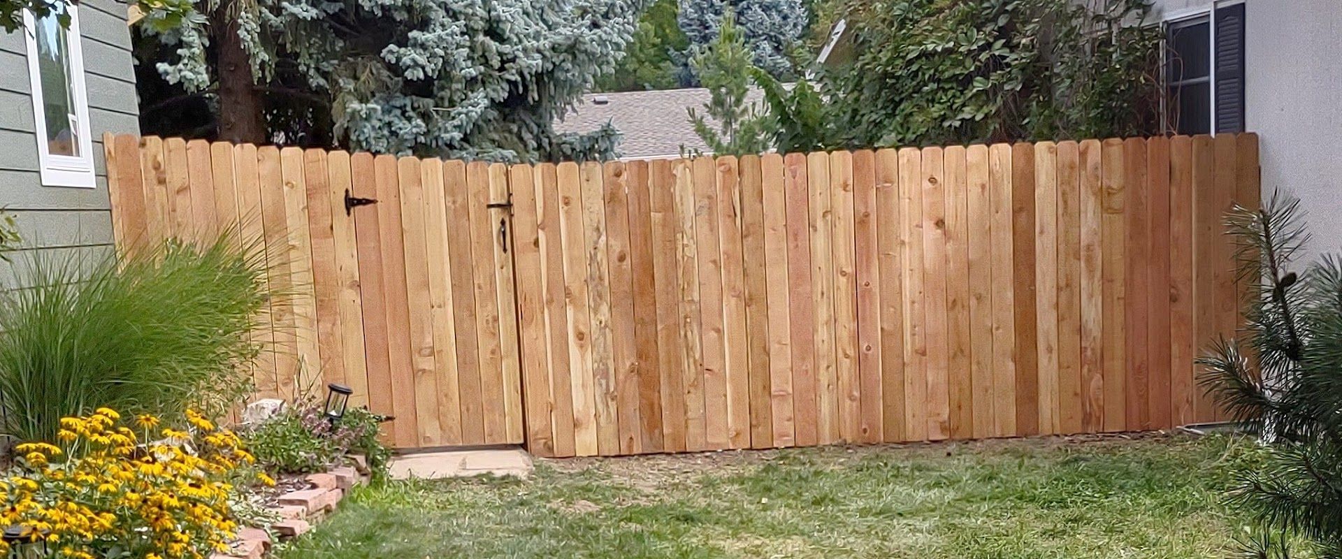 A wooden privacy fence with an integrated gate stands in a residential yard with grass and flowers in the foreground.