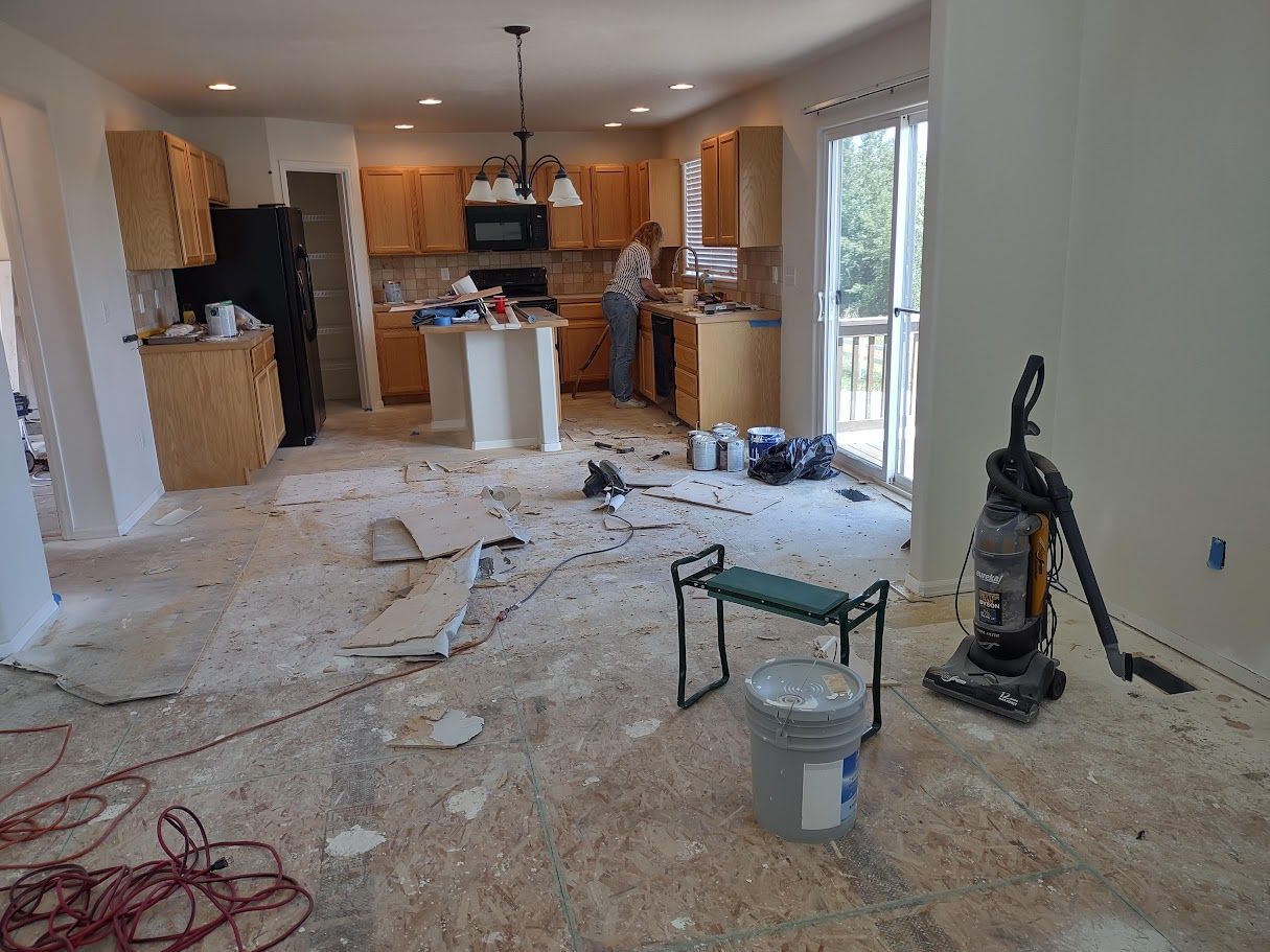 A kitchen during a renovation, with debris on the subfloor, a vacuum, and a bucket, as a person works in the background.