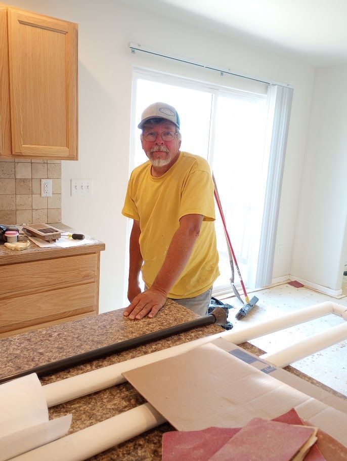 A person in a yellow t-shirt and cap stands in a kitchen with tan cabinets and a countertop during a home renovation.