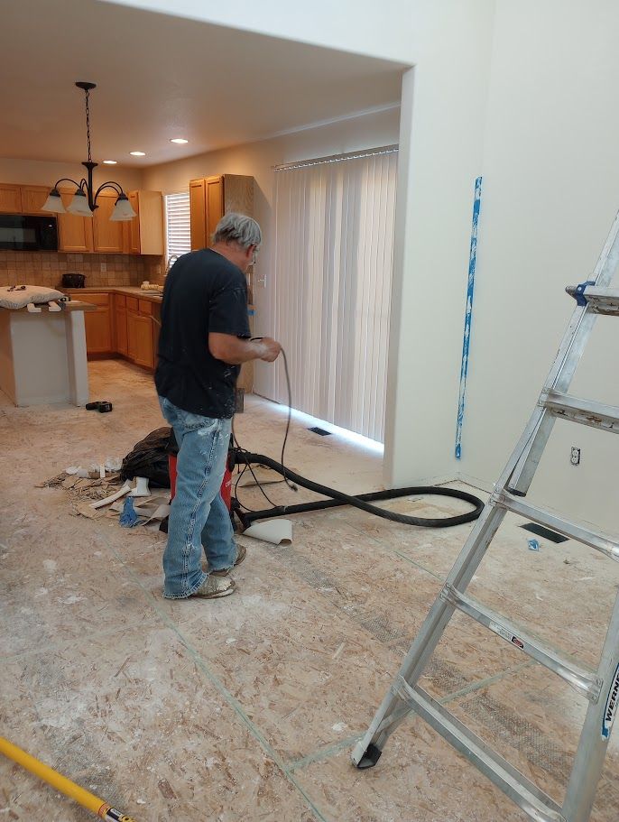 A person using a shop vacuum to clean a floor in an unfinished kitchen renovation with a ladder in the foreground.