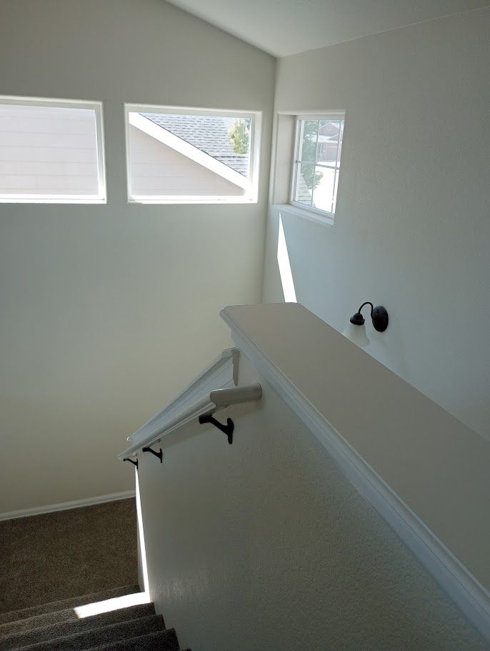 A staircase with a beige handrail and carpet, leading up toward three windows and a wall-mounted light fixture.