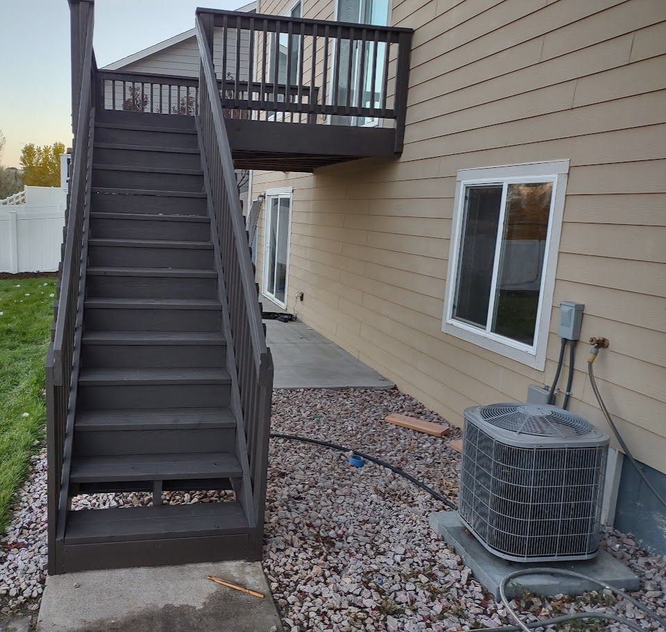 Dark wooden stairs lead up to a second-story deck next to a beige house with an air conditioning unit on a gravel yard.