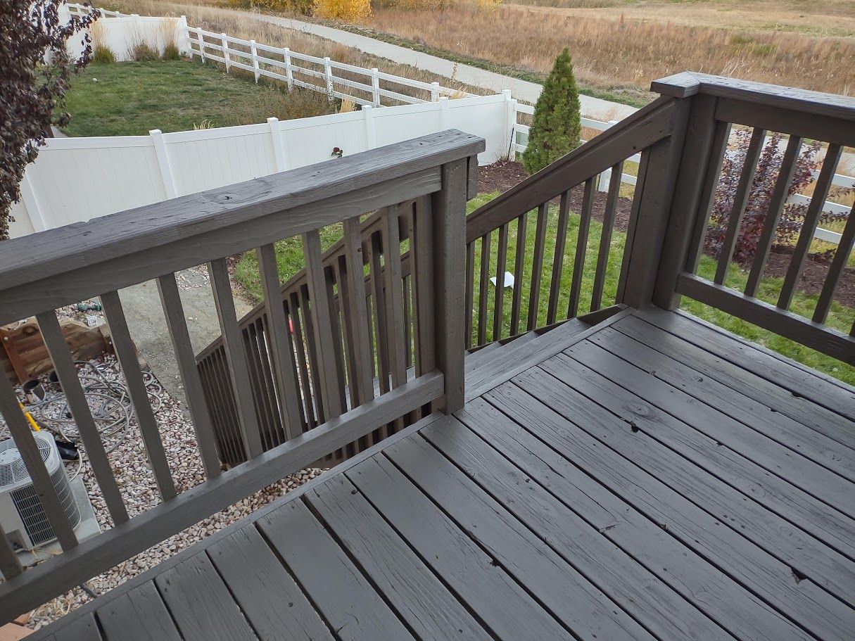 A wooden deck with a dark gray stain, featuring a railing overlooking a backyard with a white fence and grass.