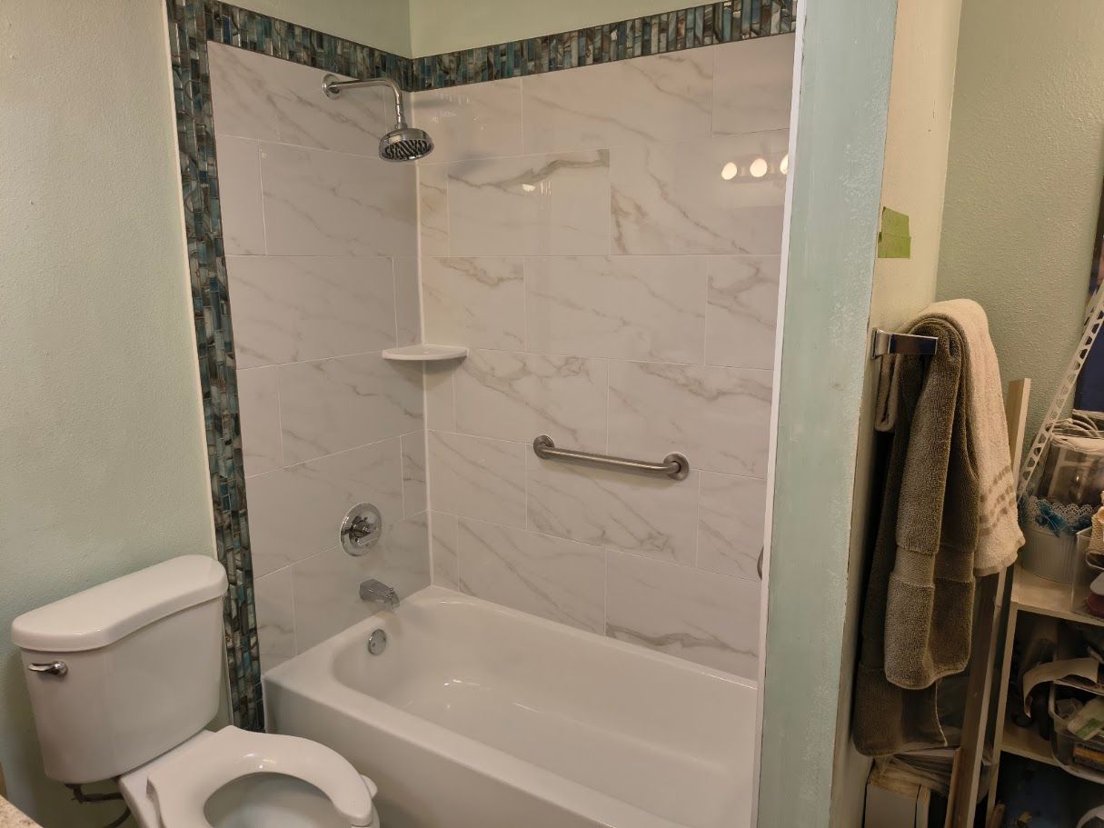 A bathroom with a white toilet and a marble-patterned tub surround featuring a showerhead and metal grab bar.