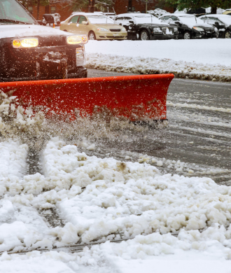 Red snowplow clearing a snowy road in winter with cars parked nearby