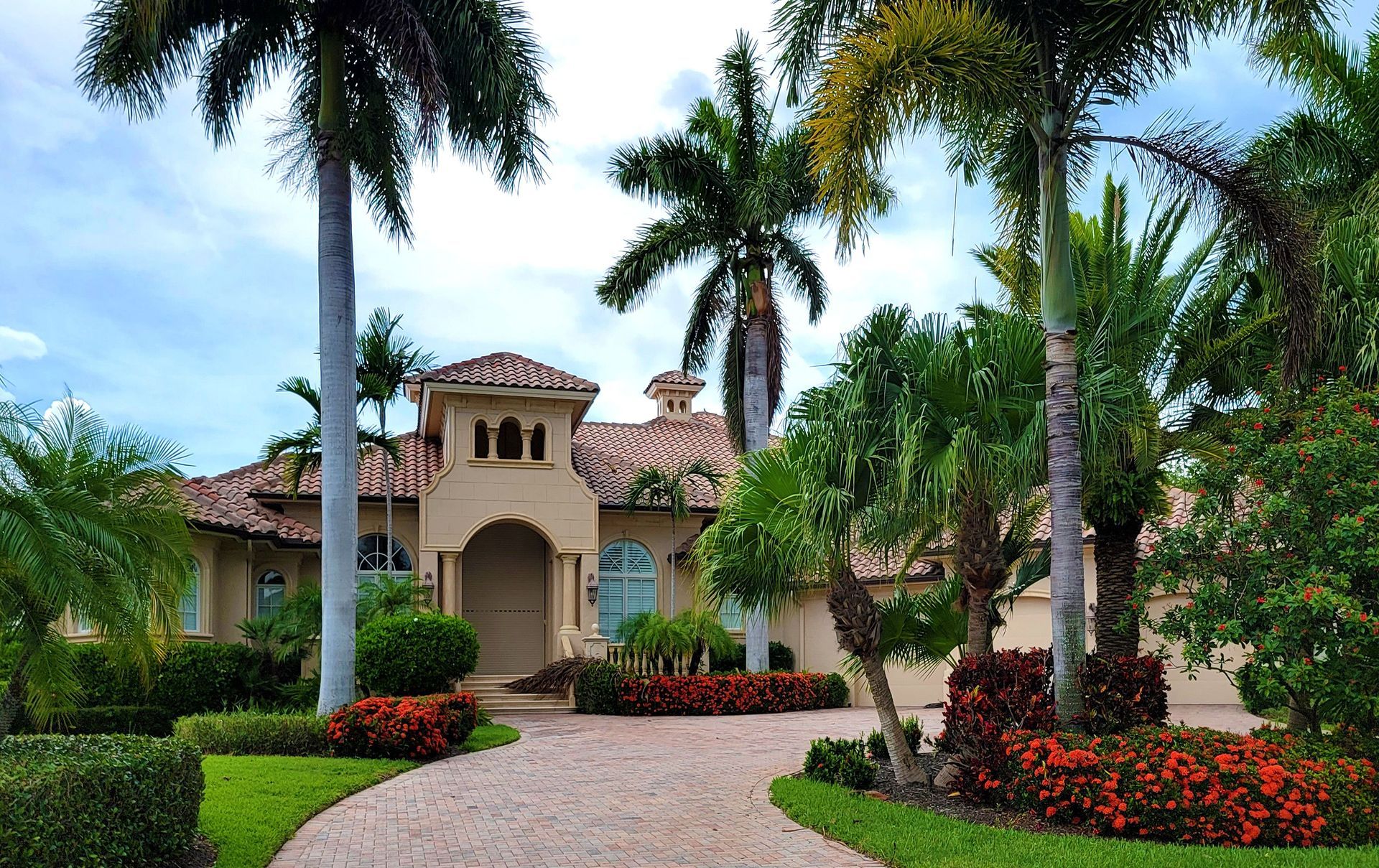 Luxurious house with palm trees, a cobblestone driveway, and red flowers in a landscaped garden