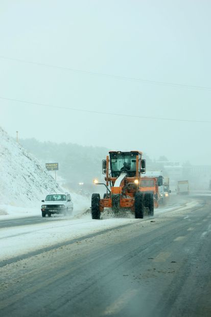 Orange snowplow clearing a snowy road beside a white car in a foggy winter scene