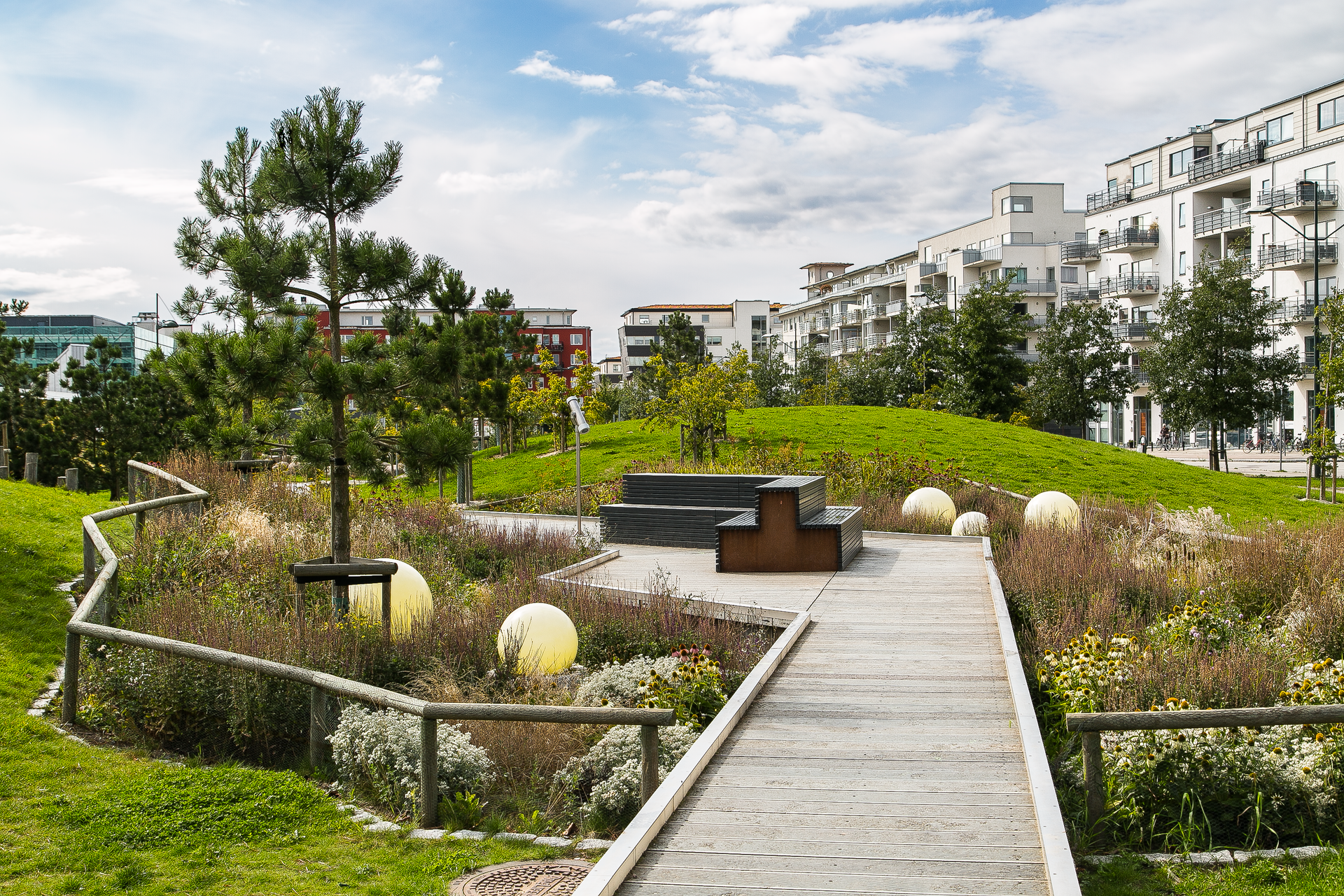 Landscaped park with a wooden walkway, flowerbeds, and modern apartment buildings under a blue sky