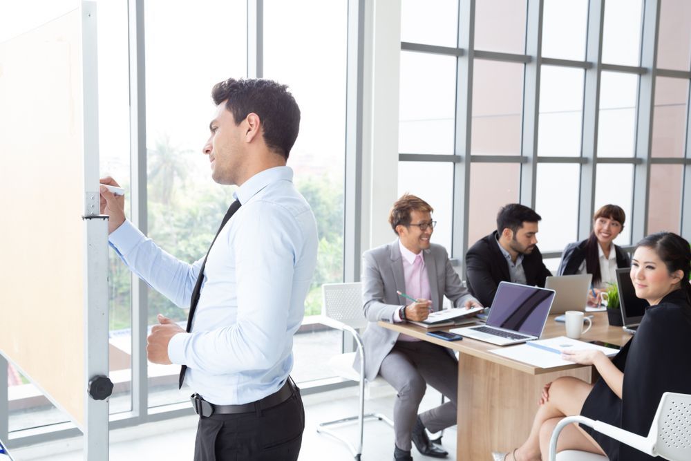 Man presenting at a whiteboard in an office meeting with colleagues seated at a table.
