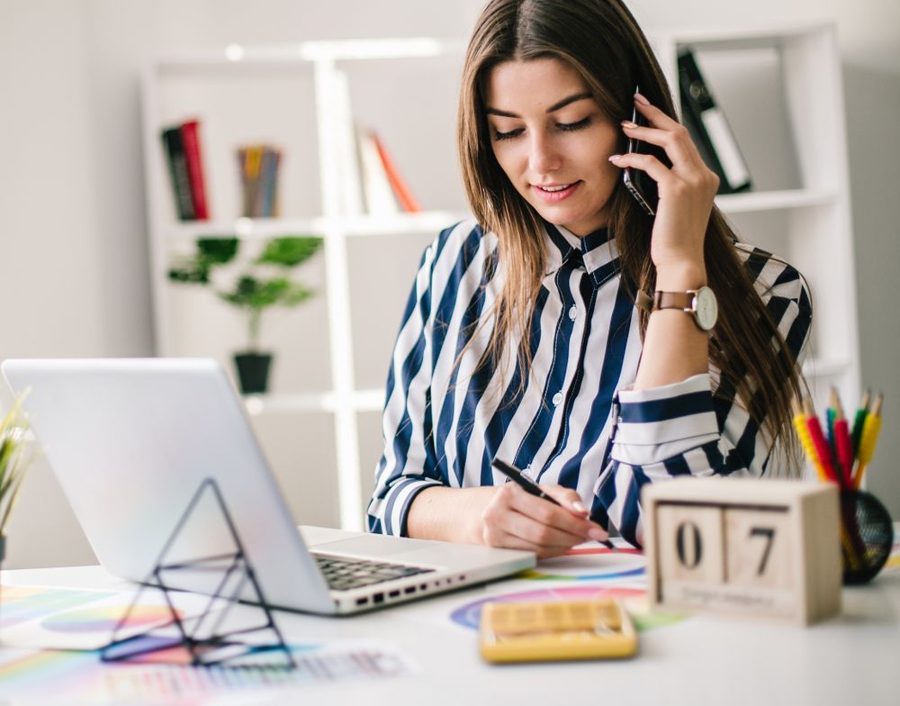 Woman working at a desk with a laptop, talking on the phone, with calendar blocks and stationery nearby