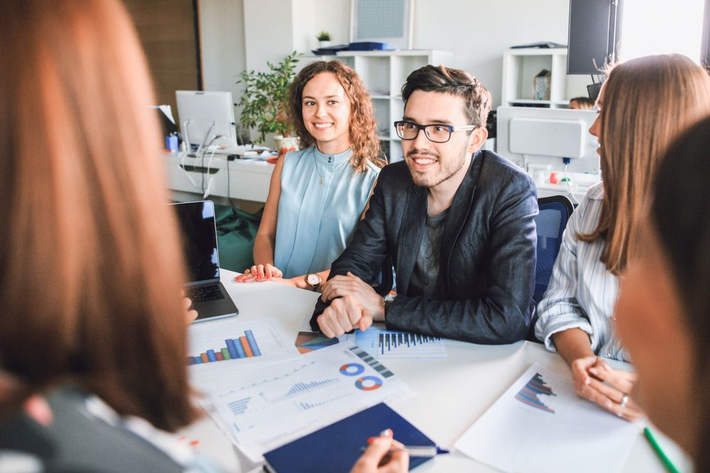 Team meeting in a bright office, with colleagues reviewing charts and smiling around a table.