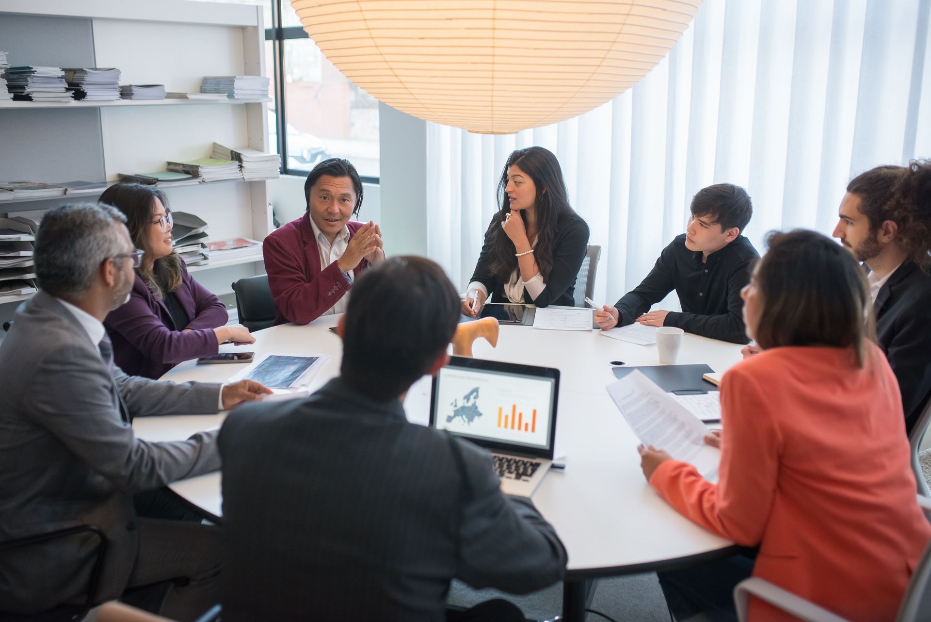 Business meeting around a conference table with laptops, papers, and a chart on screen