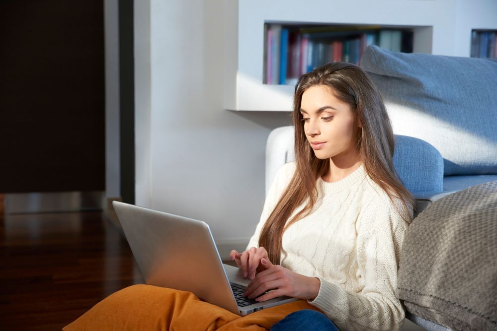 Woman using a laptop while sitting on the floor with a blanket; sunlight streams in.