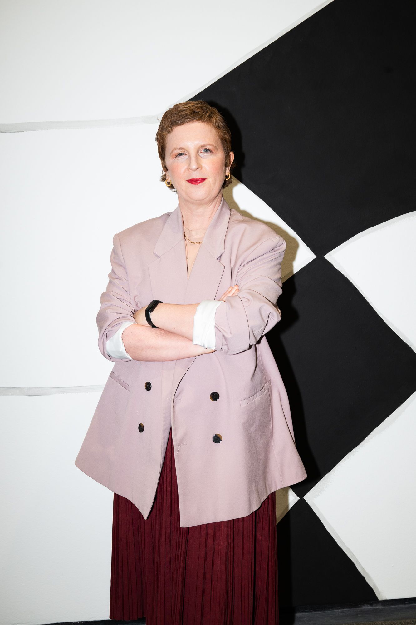 Woman in pink blazer and burgundy skirt, arms crossed, stands before black and white geometric wall.