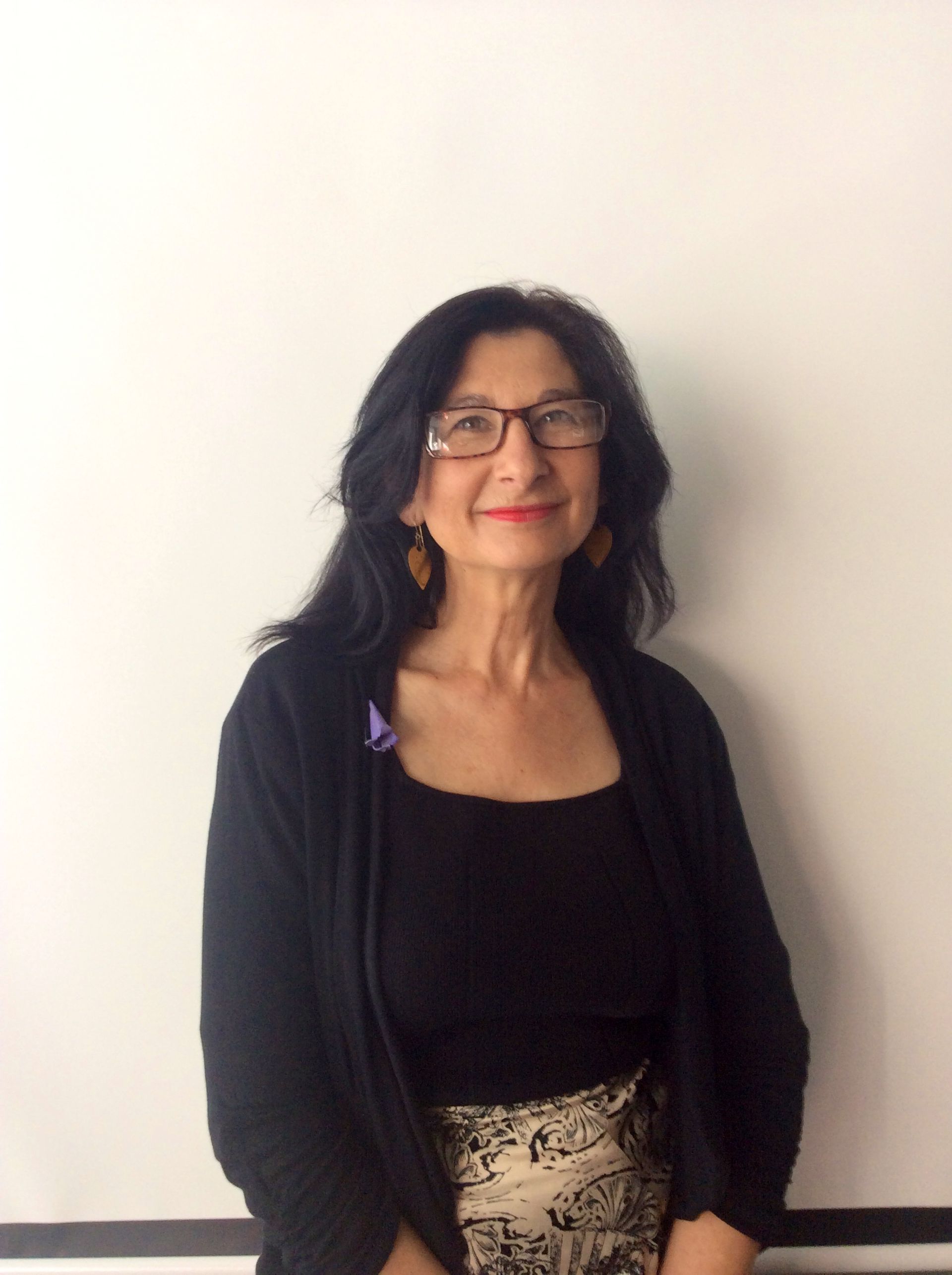 Woman wearing glasses and black cardigan, standing in front of a white wall.