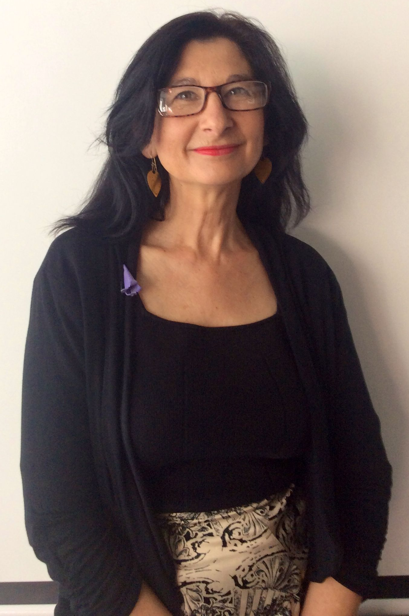 Woman with glasses, black top and cardigan, printed skirt, standing against a white wall.